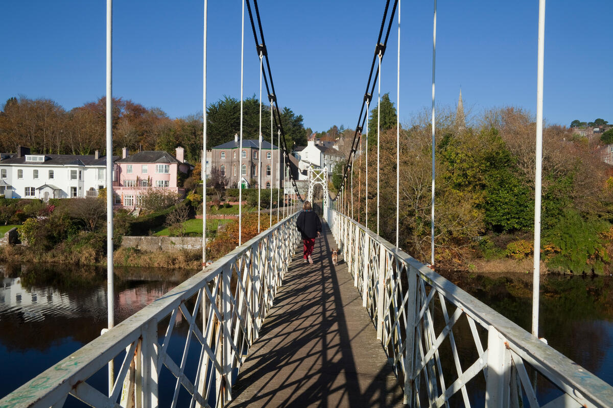 A person walking across Daly&rsquo;s Bridge over the River Lee, with houses and trees along the riverbank in Cork City.